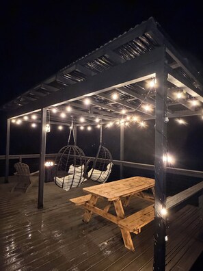 Un espace extérieur abrité, doté d’une table en bois et de deux fauteuils suspendus, éclairé par des guirlandes lumineuses.