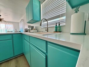 Eine Küche mit türkis-blauen Schränken, einer weißen Arbeitsplatte, einem Fenster mit Jalousien und einem Deckenventilator.