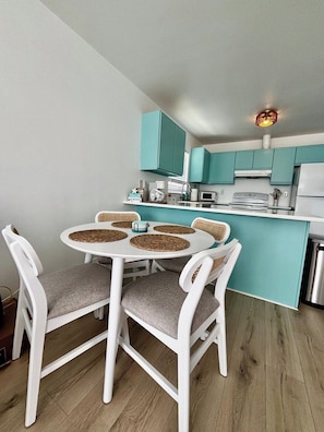 Una cucina compatta con mobili color petrolio, un tavolo da pranzo bianco con sottopiatti intrecciati e un moderno lampadario.
