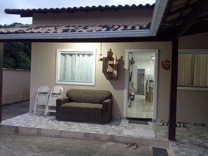 A covered outdoor area with a sofa, a window with curtains, a door leading to a kitchen, and a small step with a mat. - New Listing (Cabo Frio)