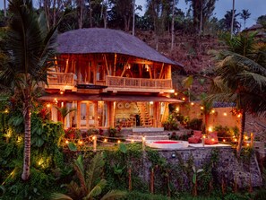 Une maison en bois à deux étages, dotée d’un balcon, entourée d’une végétation luxuriante et illuminée de guirlandes lumineuses.