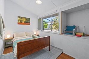 Un dormitorio con cama de madera, una mesita de noche con lámpara, una ventana con vistas al exterior y un pequeño estante con juguetes.