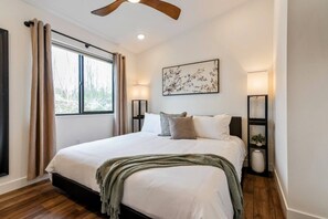 Un dormitorio con cama, ventilador de techo, ventana con cortinas, lámpara de pared y un cuadro enmarcado.