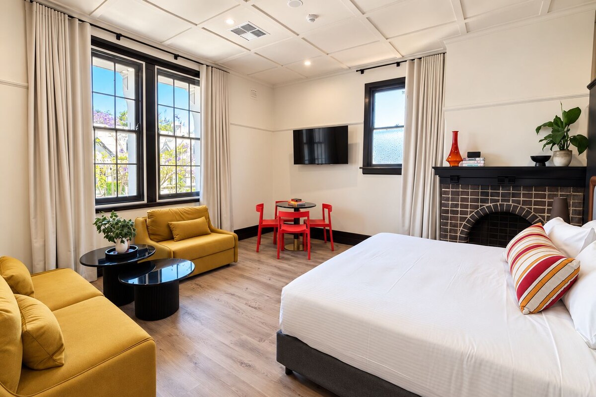 A hotel room with a bed, a sofa, a small table, a fireplace, and a TV.