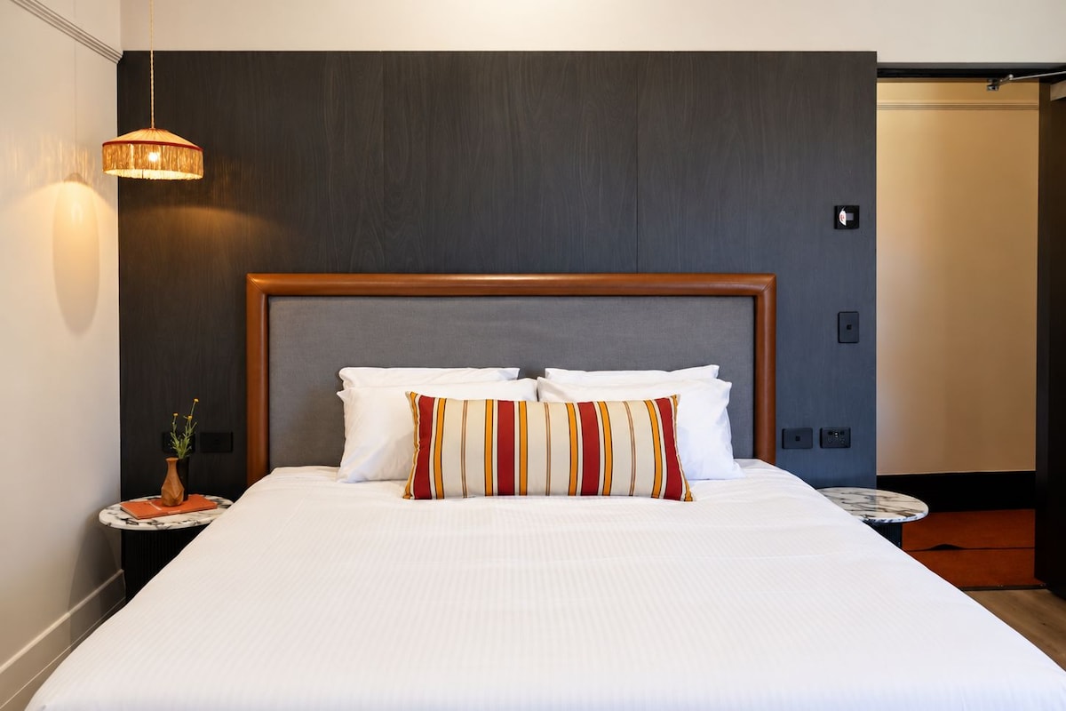 A hotel room with a large bed, a wooden headboard, a bedside table with a vase, and a hanging light fixture.