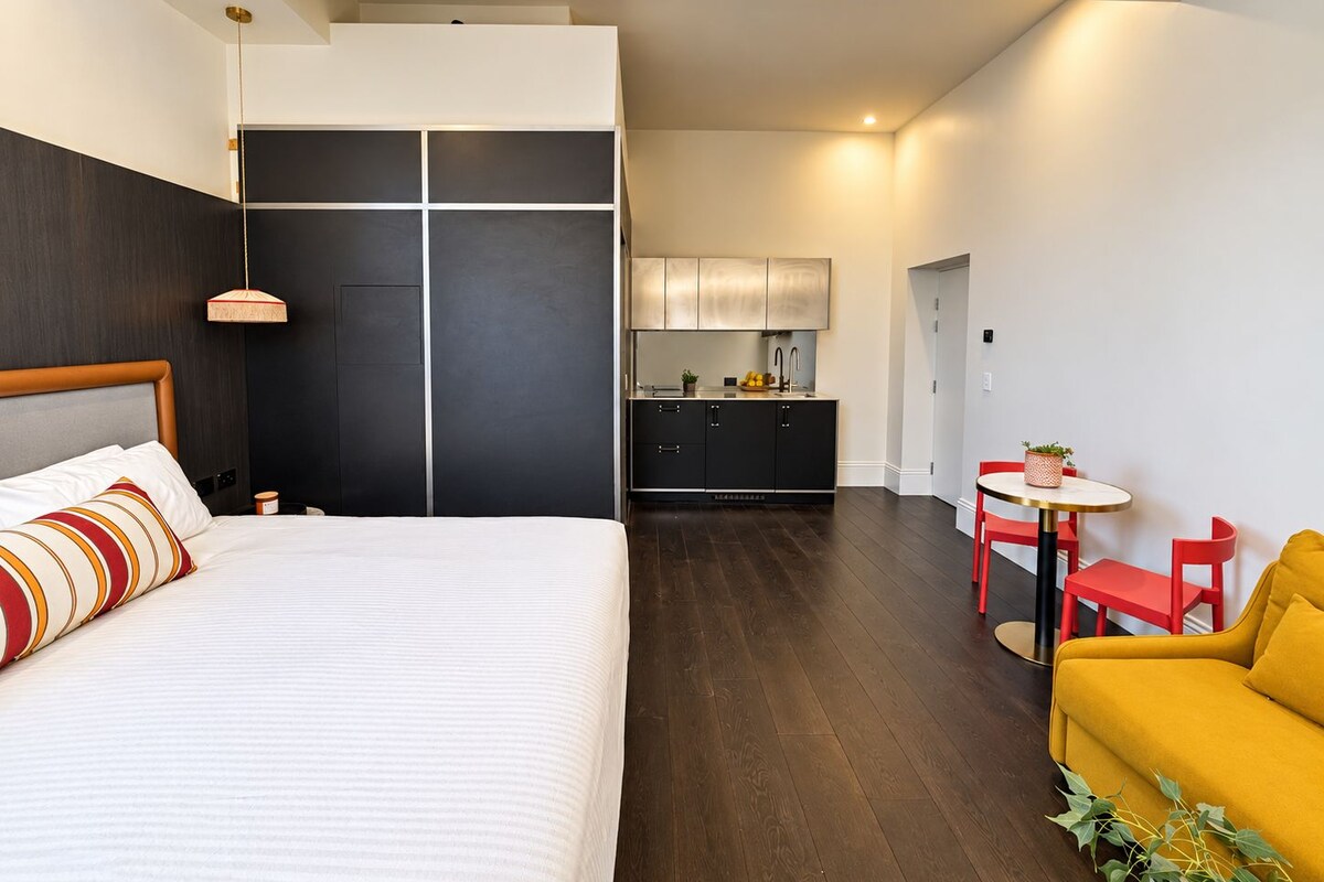 A modern hotel room with a large bed, a small kitchen area, and a dining table with chairs.
