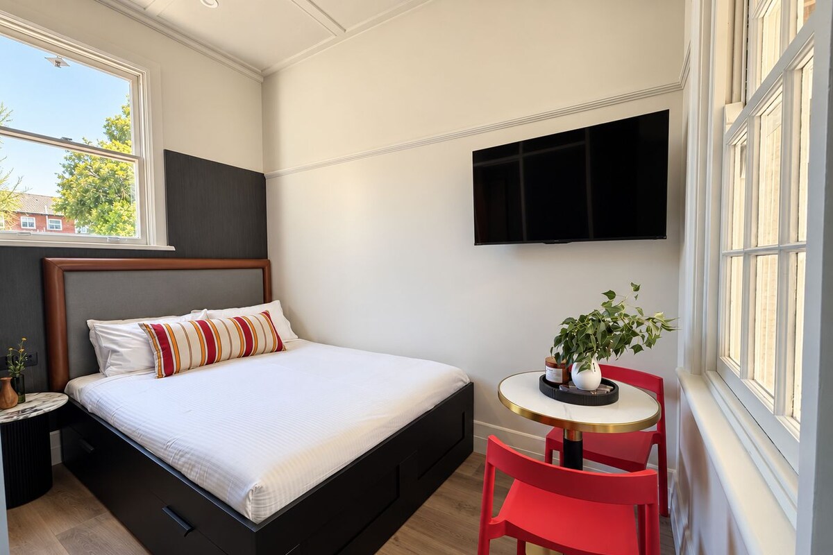 A bedroom with a bed, a bedside table, a red chair, a small table with a plant, and a TV mounted on the wall.