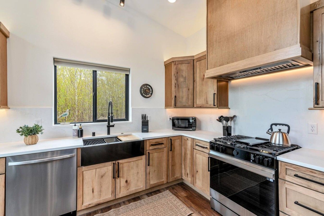 A modern kitchen with wooden cabinets, a stainless steel dishwasher, a built-in oven, and a window with a view of trees.