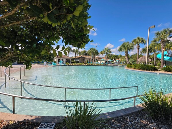 A clear swimming pool with a no entry sign, surrounded by palm trees and a building with a tiled roof.