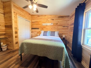 Quarto com painéis de madeira, uma cama, um ventilador de teto, uma janela com cortinas e um aviso na parede.