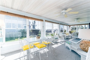 A sunlit room with a glass table, white wicker furniture, and a view of a house through large windows. - Weekapaug's Largest Rental — 7 Bedrooms, Sleeps 14, Steps to Everything (Westerly)