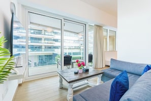 A modern living room with a large window, a sofa, a coffee table, and a view of a building complex. - Luxury Jr. 1BR in Mayan Yas Island (Abu Dhabi)
