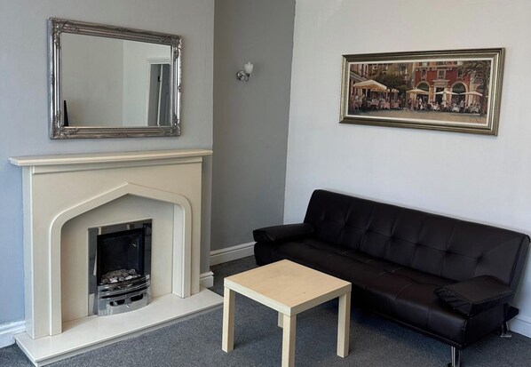 A living room with a fireplace, a black leather sofa, a wooden coffee table, a framed picture on the wall, and a mirror above the fireplace. - 2 Bed House Sleeps 3 - Garden - Pets Welcome (Middlesbrough)