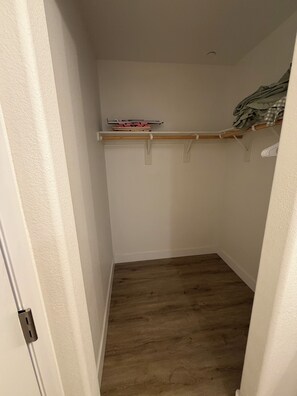 Ein schmaler Flur mit Holzboden, einer weißen Tür links und einer weißen Wand mit einem Regal, das Bücher und einen kleinen Gegenstand enthält.