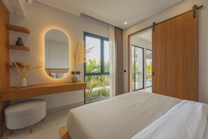 Un dormitorio con una cama grande, una mesita de noche con un florero, un espejo y un ventanal corredera que da a un balcón con plantas.