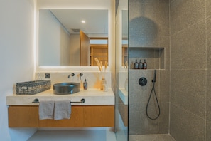 Un baño moderno con un espejo grande, un lavabo redondo, armarios de madera y una zona de ducha con mampara de vidrio.