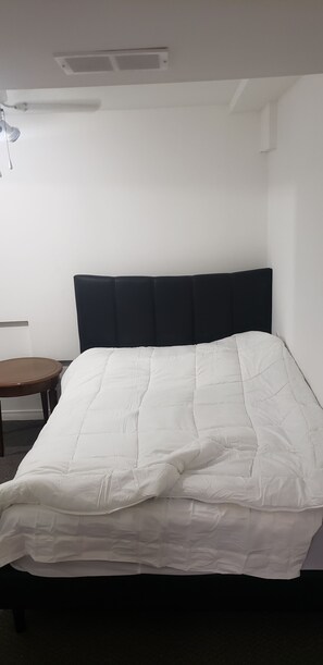 Yhden hengen sänky, jossa on valkoinen peitto, tumma pääty ja pieni puinen yöpöytä sen vieressä.