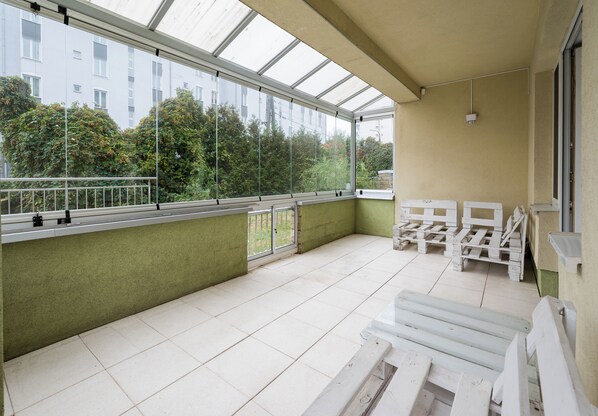 Un balcon donnant sur un immeuble et des arbres, avec une rambarde en métal et des meubles blancs.