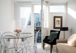 Una sala de estar moderna con una mesa de comedor de vidrio, sillas transparentes, un sillón de terciopelo negro, un sofá beige y una obra de arte enmarcada en la pared.