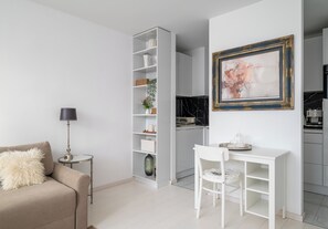 A modern living room with a sofa, a small side table, a floor lamp, a white dining table with chairs, a white shelving unit with decorative items, and a kitchen area with a marble countertop. - Grzybowska 14th Floor Royal Apartment (Warszawa)