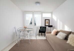 Una sala de estar moderna con una mesa redonda de vidrio, sillas traslúcidas, un sillón negro de terciopelo con un cojín mullido, un sofá marrón con cojines suaves y un cuadro en la pared.