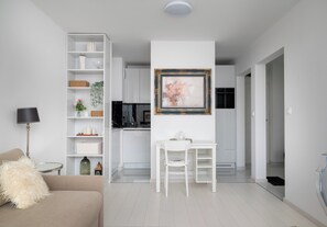 Un salon moderne avec un canapé, une table à manger, un meuble blanc avec des étagères et, au fond, un coin cuisine.