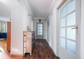 Un couloir avec un plancher en bois, une porte blanche avec des panneaux de verre et un mur bleu avec une fenêtre.