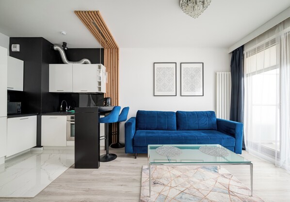 Una sala de estar moderna con un sofá azul, una mesa de centro de vidrio y una cocina con armarios blancos y encimera negra.