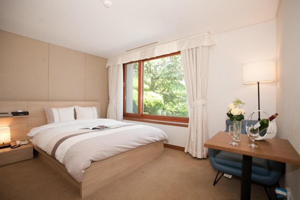 A hotel room with a bed, bedside table, lamp, and a view of greenery outside.