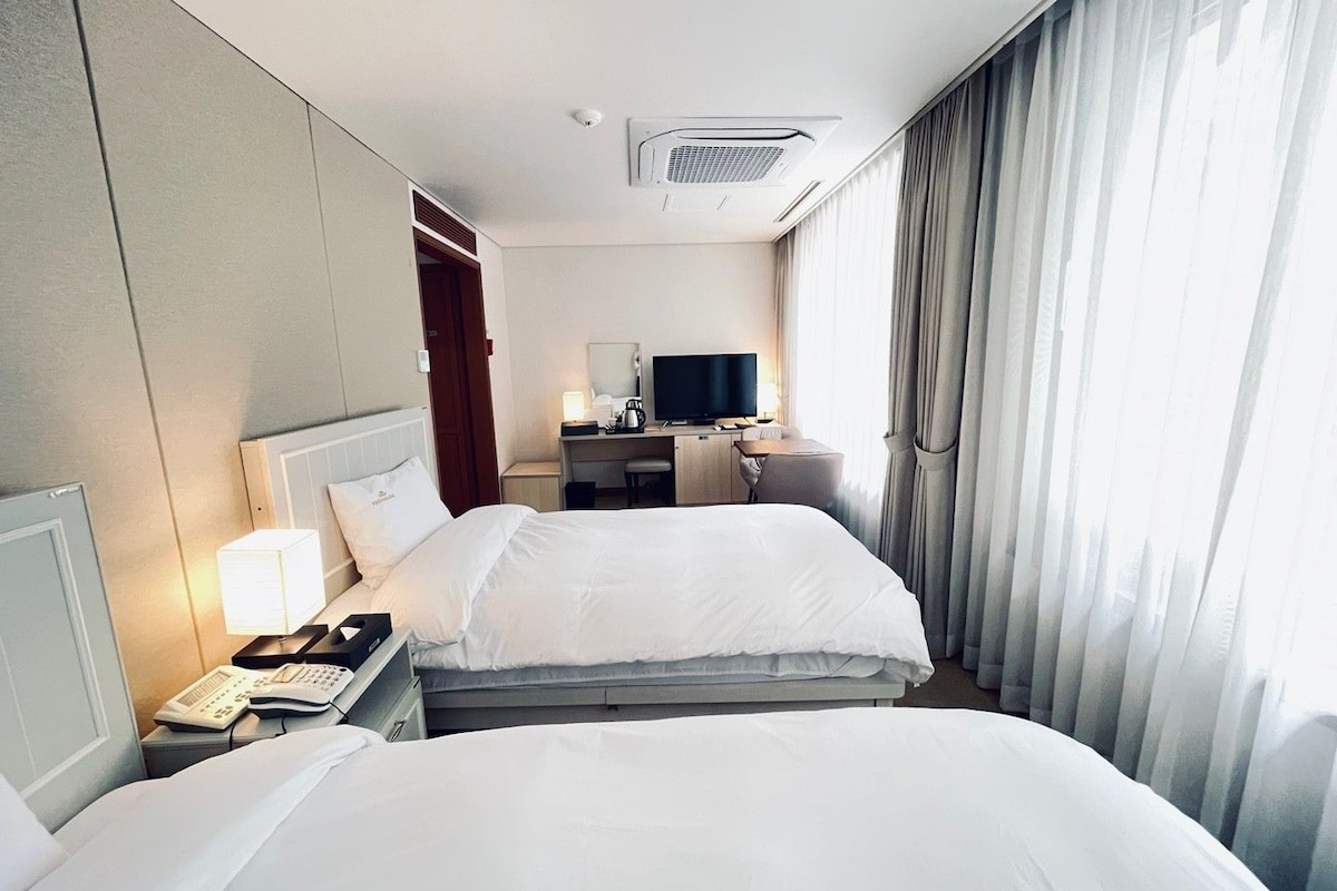 A hotel room with two beds, a desk with a television, a phone, and a window with curtains.