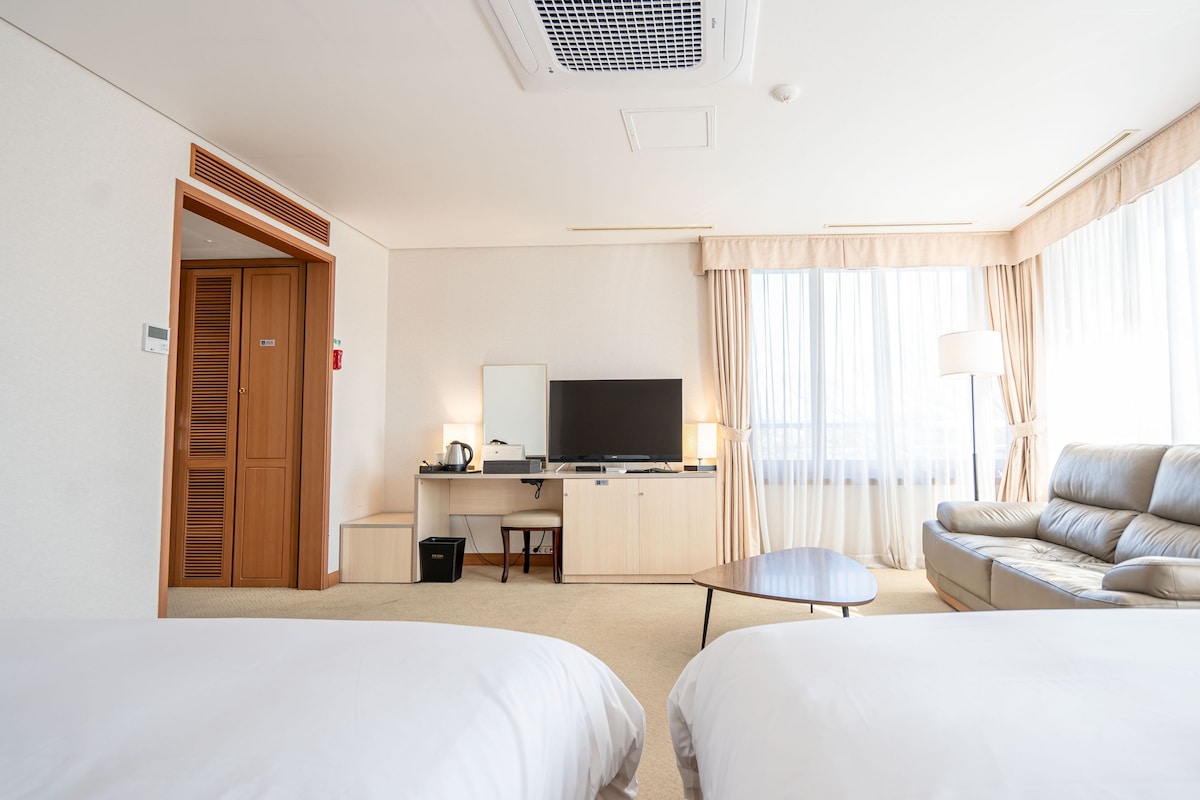 A hotel room with two beds, a desk with a TV, a sofa, and a window with curtains.