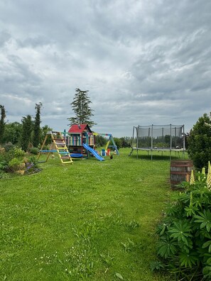 Un parc infantil amb tobogan i cadiretes, envoltat d'herba i arbres.