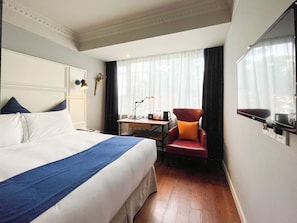 Quarto de hotel com cama, escrivaninha, cadeira e televisão.