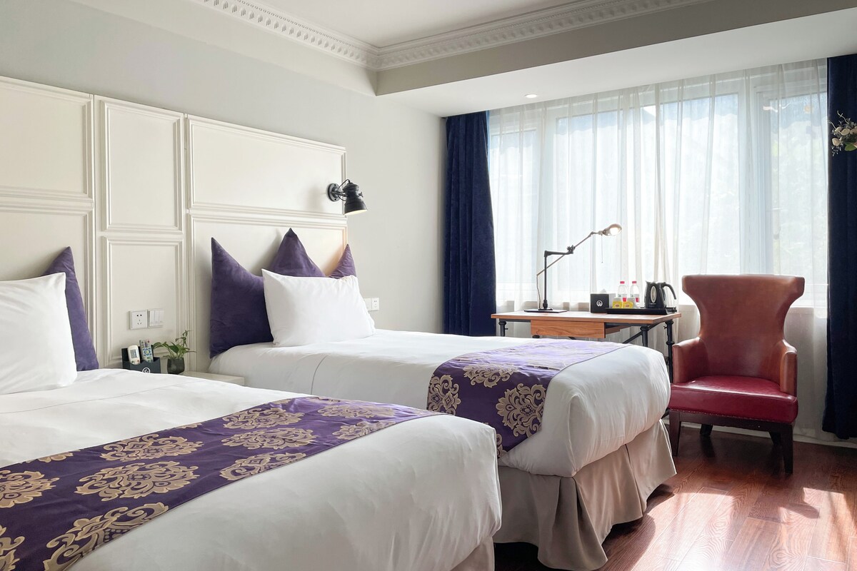 Quarto de hotel com duas camas, uma escrivaninha com luminária, uma cadeira e uma janela com cortinas.