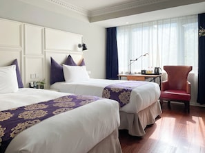 Quarto de hotel com duas camas, uma escrivaninha com luminária, uma cadeira e uma janela com cortinas.