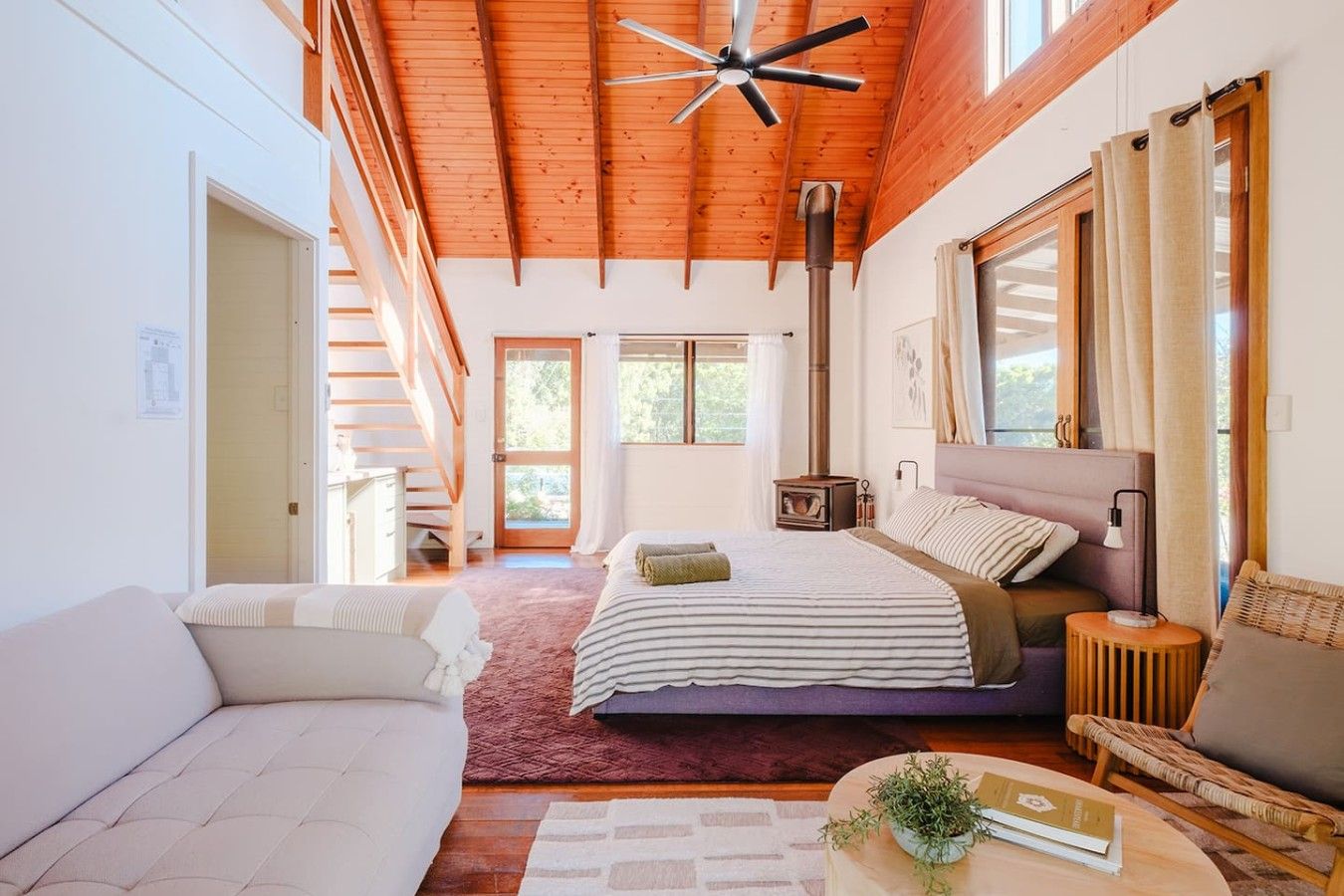 A spacious bedroom with a large bed, a wooden ceiling, a built-in wood stove, and a view of the outdoors.