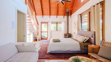 A spacious bedroom with a large bed, a wooden ceiling, a built-in wood stove, and a view of the outdoors.
