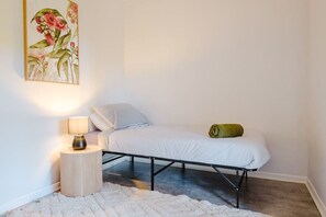 Una piccola camera con un letto singolo, una struttura in metallo, una lampada da comodino, un asciugamano arrotolato e una stampa botanica incorniciata al muro.