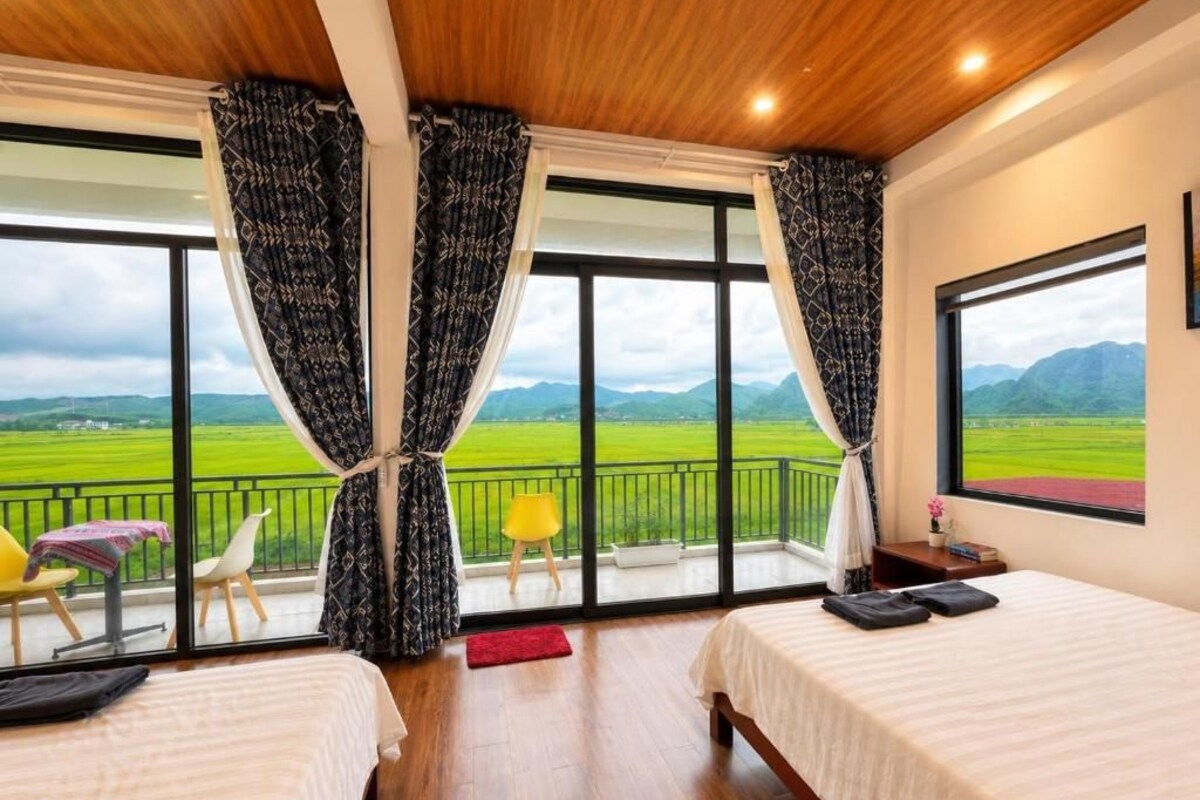 Habitación de hotel con dos camas, un balcón con vistas al campo y un amplio ventanal con cortinas.