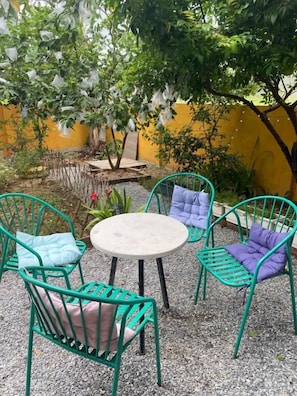 Un espacio de estar al aire libre con sillas de metal verde y una mesa redonda, rodeado de plantas y una pared amarilla.