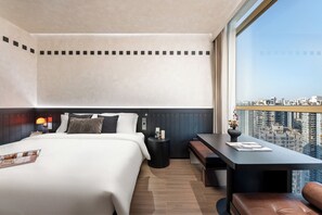 Quarto de hotel moderno com uma cama grande, uma mesinha e um banco. Vista da cidade visível através de janelas amplas.