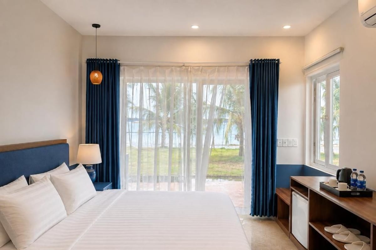 A hotel room with a bed, a desk, a lamp, a view of palm trees, and a window with curtains.