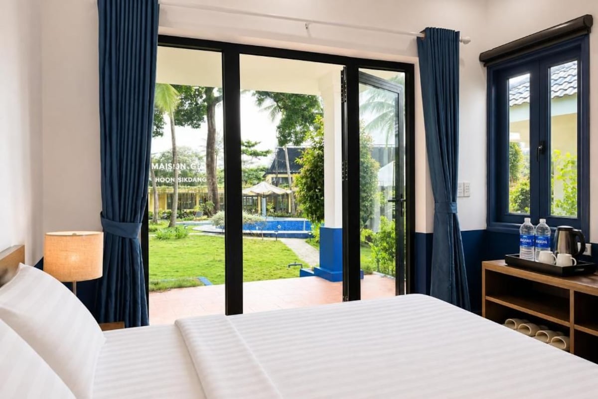 A hotel room with a bed, a nightstand, a lamp, a table with bottles and a tray, and a view of a pool and garden through a glass door.