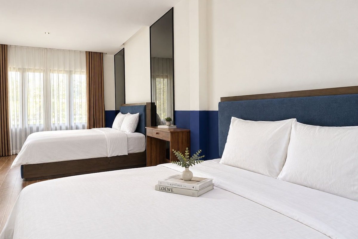 A hotel room with two beds, a bedside table with a lamp, a mirror, and a window with curtains.