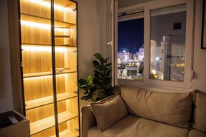 A room with a wooden shelving unit, a window with a view of a cityscape at night, and a beige sofa. - STOUNE BRIDGE APARTMENT 24 (Skopje)