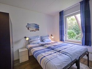 Une chambre à coucher comprenant un lit, des tables de chevet, une lampe fixée au mur, une fenêtre avec des rideaux et une vue partielle sur une porte.