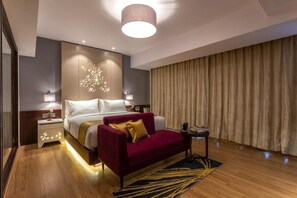 Quarto de hotel com uma cama, um sofá vermelho, uma mesa pequena, um abajur e uma cortina.