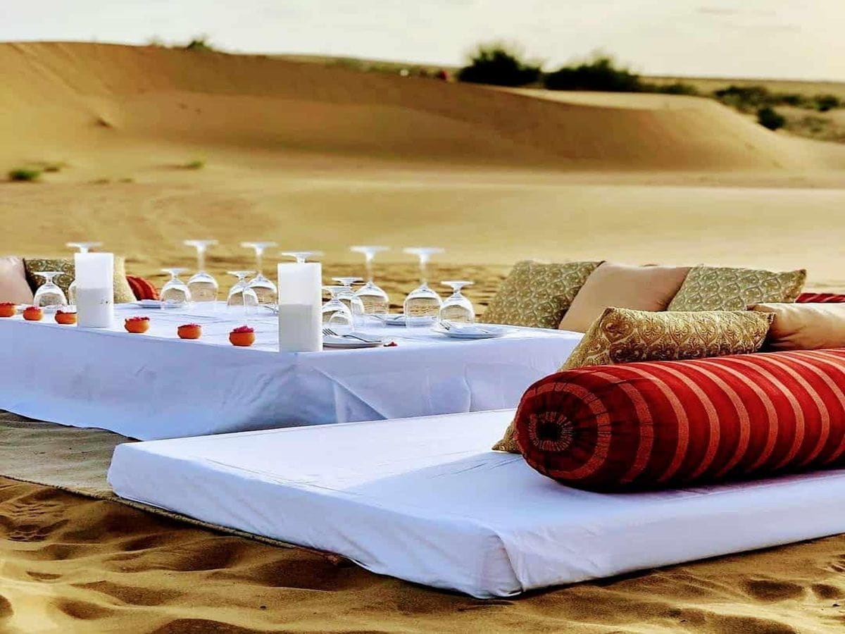 Un décor désertique avec une table dressée pour un repas, comprenant des bougies, des verres et un dessert.