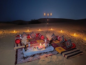 Un décor désertique avec une table dressée pour un repas, entourée de bougies et de coussins décoratifs.