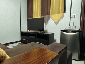A compact room with a flat-screen TV on a black stand, a small refrigerator, and a sofa with a wooden accent. - Teva's Tahiti Stay (Faaa)